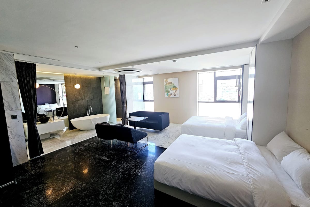 A modern hotel room with a large bed, a sitting area with a chair, a bathtub, and a large window providing natural light.