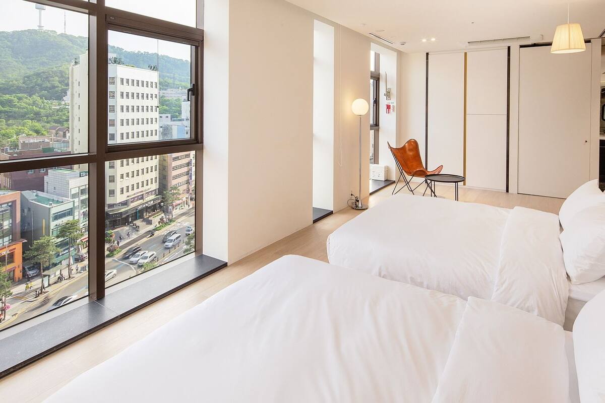 A modern hotel room with a large window offering a city view, a bed with white bedding, and a small table with a chair.