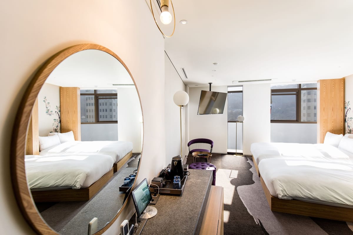 A modern hotel room with two beds, a large mirror, a flat-screen TV, a desk with a computer, and a coffee machine.