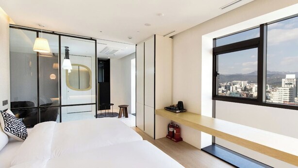 Una habitación de hotel moderna con una cama grande, un escritorio, una silla, un espejo y vistas de la ciudad a través de amplios ventanales.