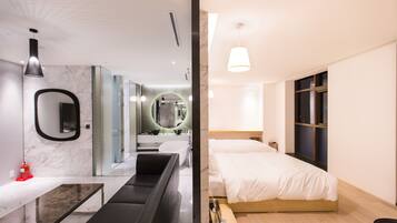 Una habitación de hotel moderna con una cama grande, una zona de estar con un banco, un baño con un espejo amplio y una mesita pequeña.