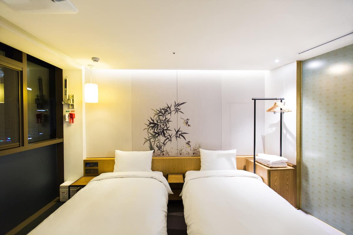 A hotel room with two beds, a bedside table, a nightstand, a wall-mounted lamp, a mirror, and a decorative bamboo plant.