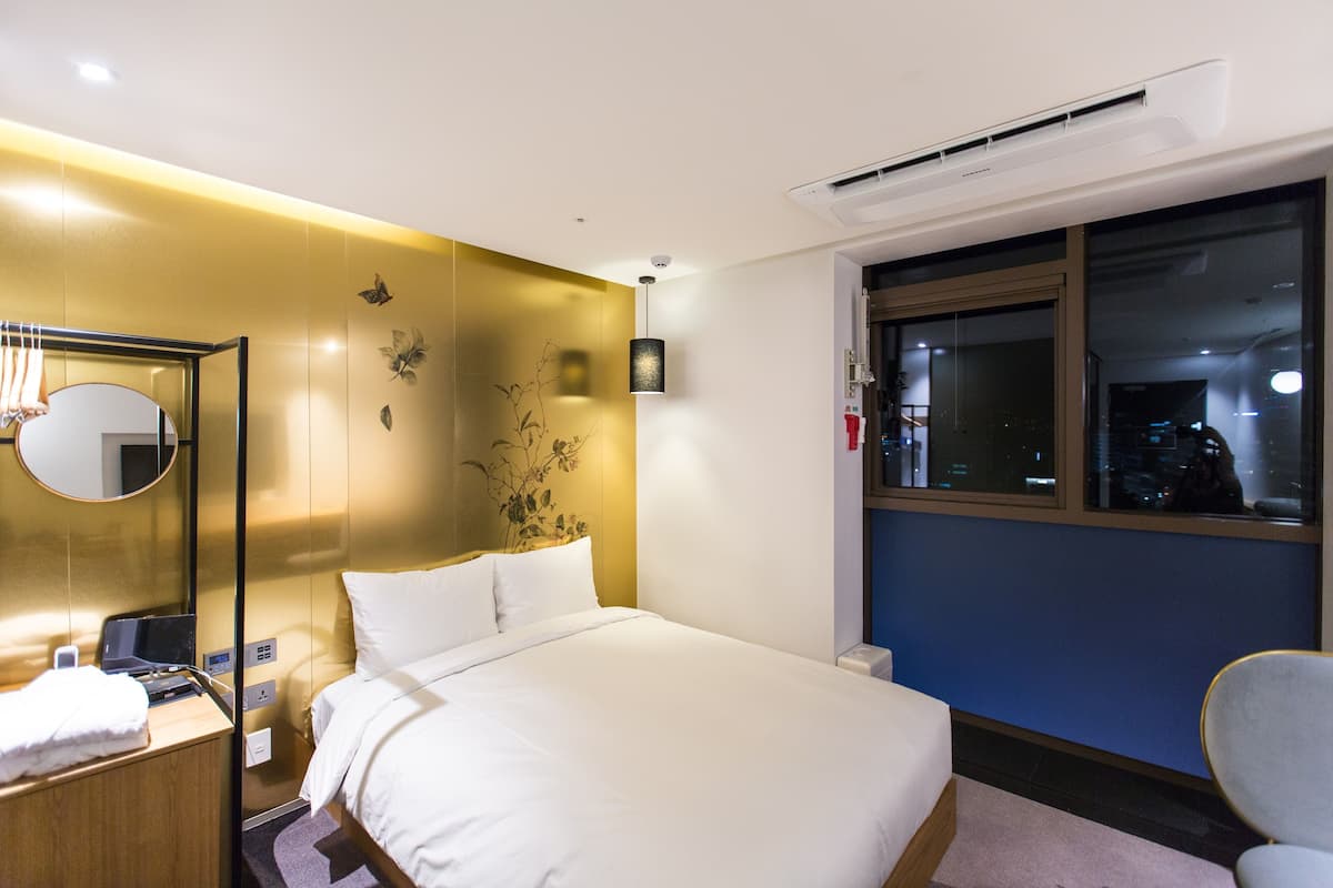 A hotel room with a bed, bedside table, lamp, and a window with a view of the city.