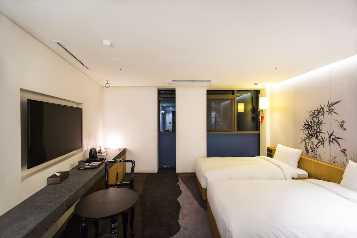 A hotel room with a large flat-screen TV, a desk with a chair, two beds, and a wall-mounted air conditioner.