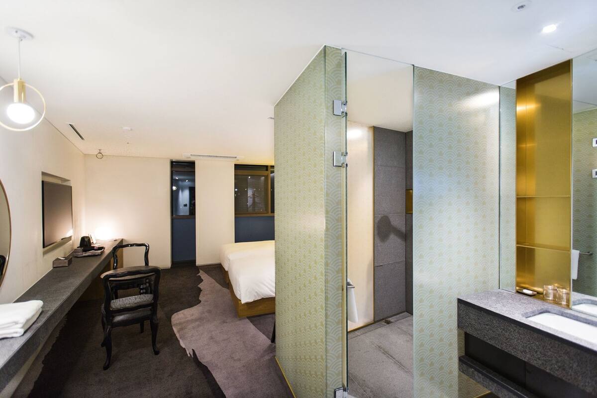 A hotel room with a bed, a desk, a chair, a mirror, and a shower area.