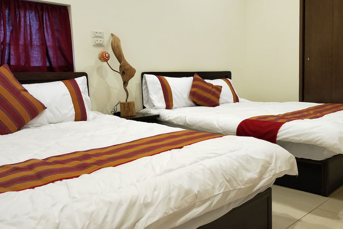 Habitación de hotel con dos camas, cada una con ropa blanca y mantas a rayas. Entre las camas hay una mesita de noche de madera con una lámpara y un objeto decorativo. La habitación tiene un diseño sencillo y limpio, con una pared lisa y una ventana con cortinas.