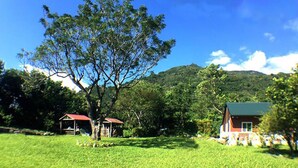 Un paisaje rural con un árbol grande, una construcción pequeña con techo rojo y un edificio más grande con techo verde.