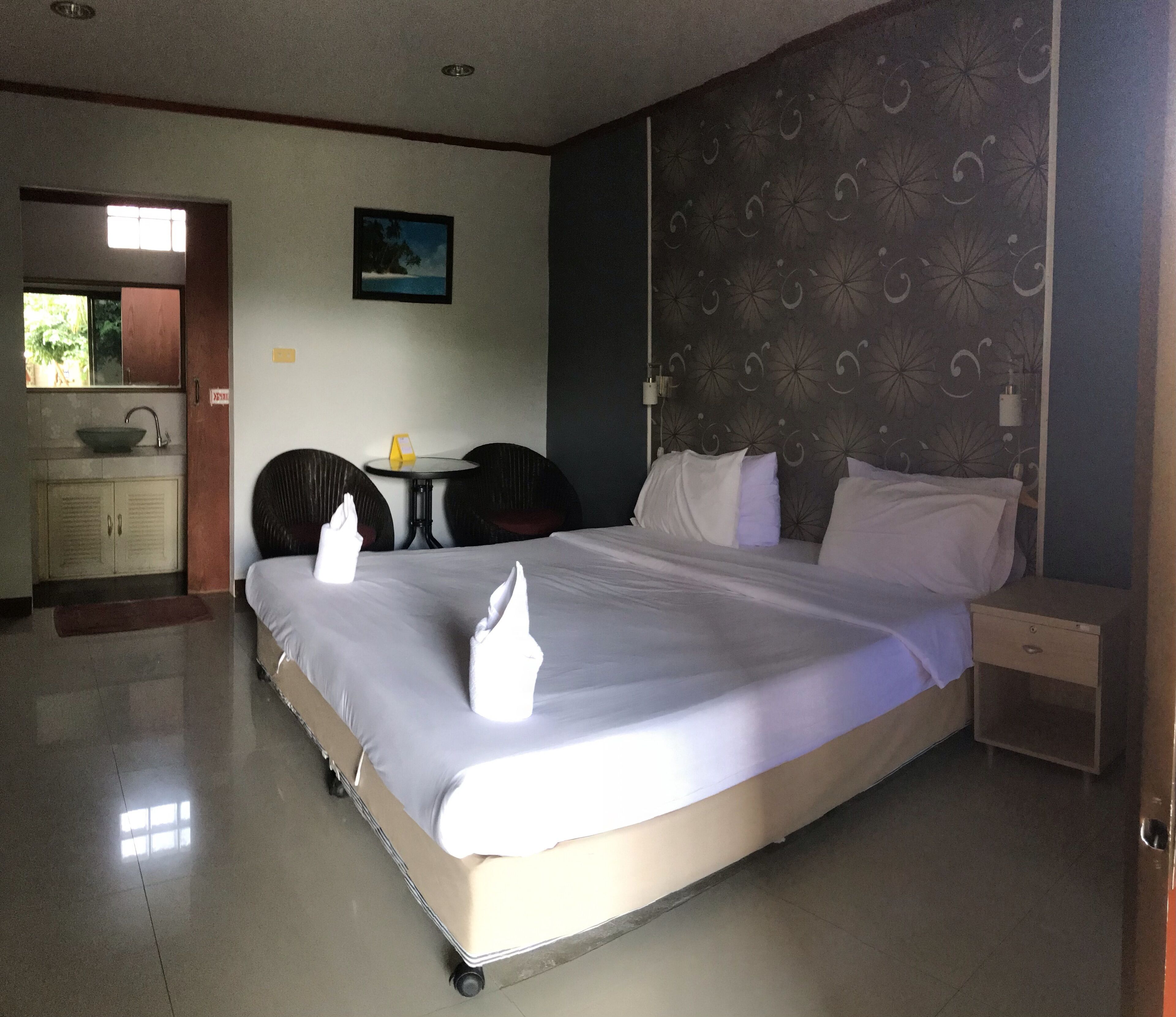 A hotel room with a large bed, a bedside table, a nightstand, a small table with a chair, a sink, and a mirror.