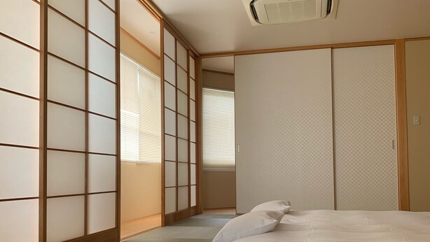 A traditional Japanese-style room with tatami flooring, a futon bed, sliding shoji doors, and a ceiling-mounted air conditioning unit. - Hotel Oxio (Okayama)