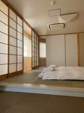 A traditional Japanese-style room with tatami flooring, a futon bed, sliding shoji doors, and a ceiling-mounted air conditioning unit. - Hotel Oxio (Okayama)