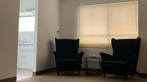 A room with two dark armchairs, a small table, a window with blinds, and a kitchenette in the background. - Hotel Oxio (Okayama)