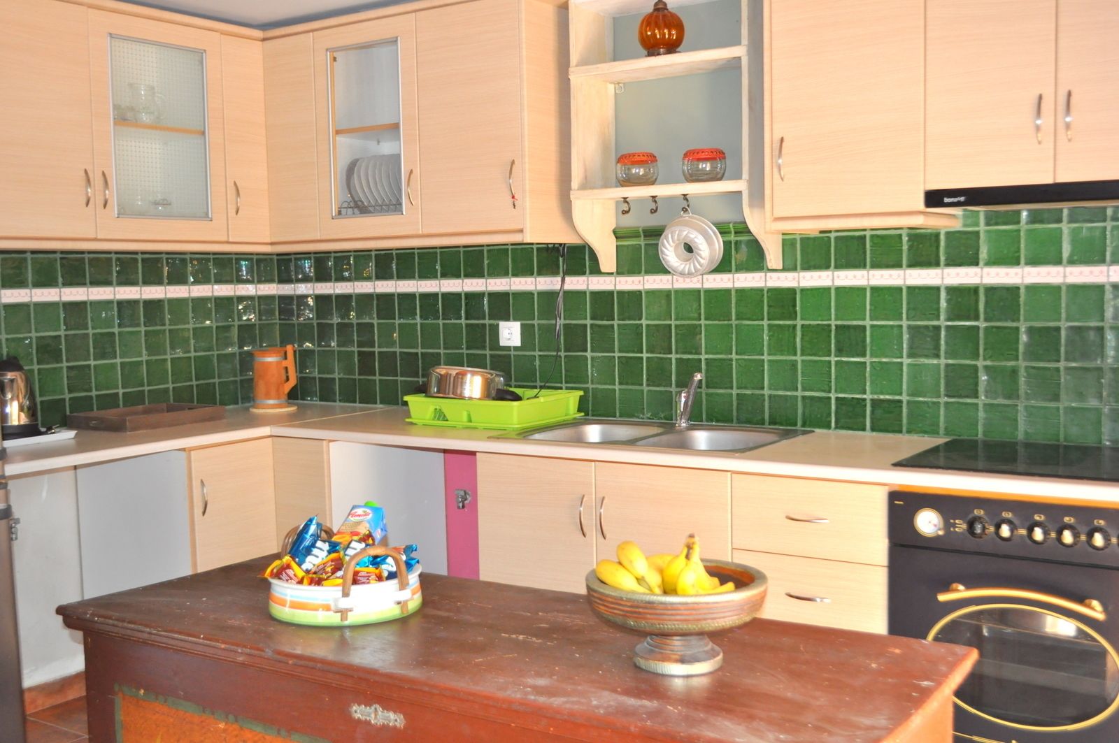 Een keuken met een groene tegelwand, houten kastjes, een fornuis en een schaal fruit op het aanrecht.