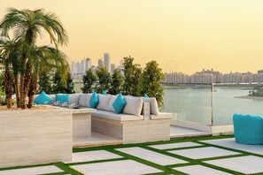 A modern outdoor seating area with a city skyline view, palm trees, and a water body. - Royal Central Hotel and Resort – The Palm (Dubai)