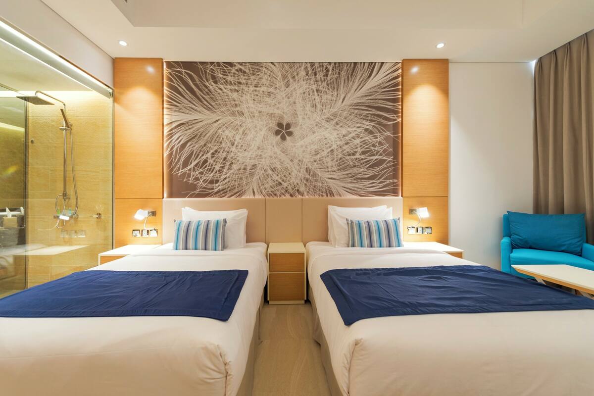 Ett hotellrum med två sängar, ett stort konstverk på väggen, en dusch och en blå fåtölj.