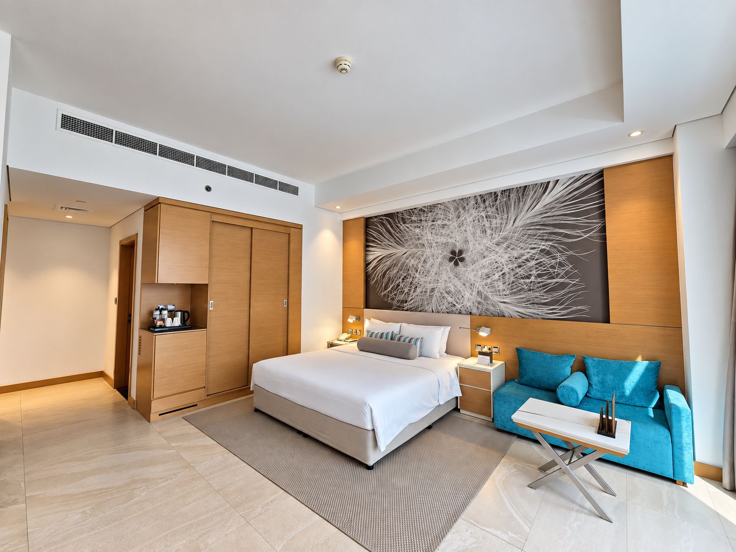 Une chambre d’hôtel moderne avec un lit, un canapé bleu, une grande œuvre d’art abstraite accrochée au mur et une armoire en bois.