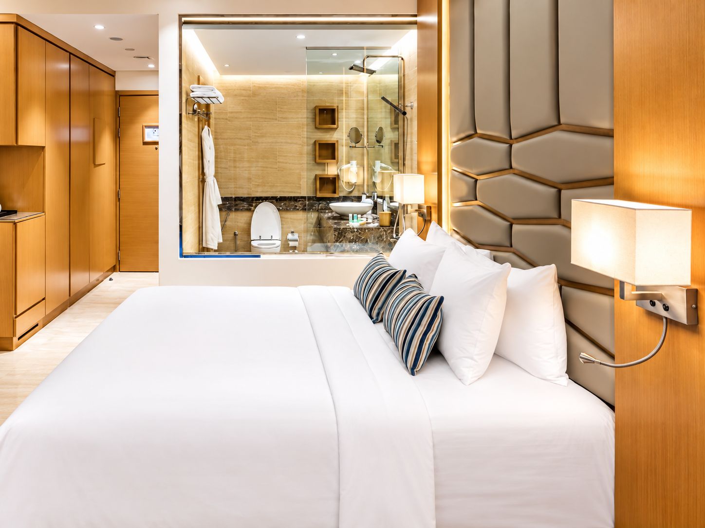 A hotel room with a large bed, a wooden wardrobe, a bathroom with a glass shower, and a view of the room.
