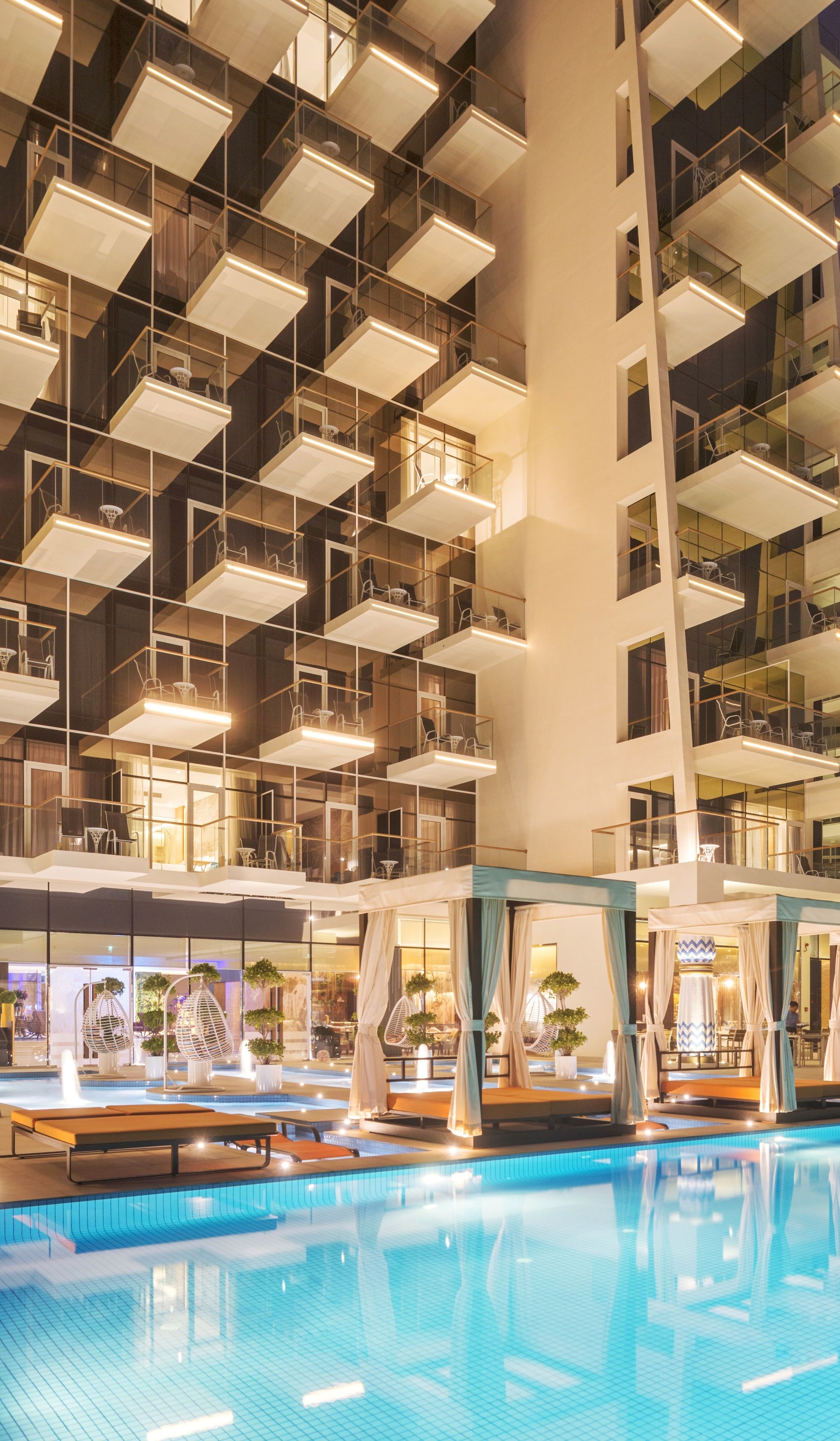 A modern hotel interior with a swimming pool, multiple balconies, and a high ceiling with geometric lighting fixtures.