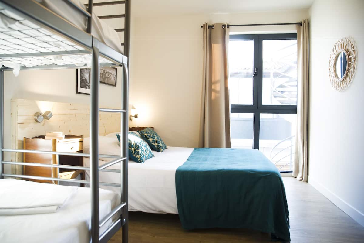 A room with bunk beds, a single bed with a blue blanket, a bedside table with a lamp, and a window with curtains.