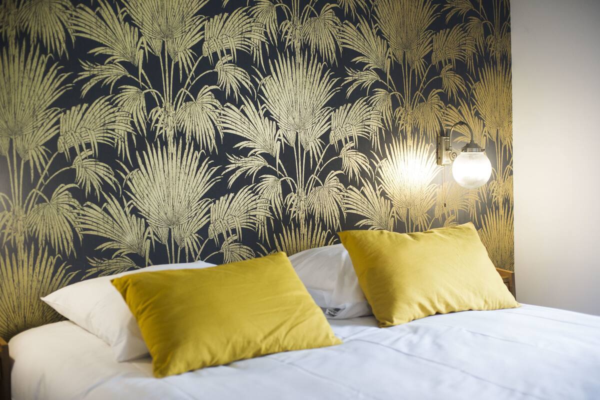 A bed with white bedding and two yellow pillows, a wall with a black and gold leaf pattern, and a wall-mounted light fixture.