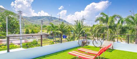 A rooftop terrace with artificial grass, two red lounge chairs, a bicycle, and a view of a mountainous landscape.