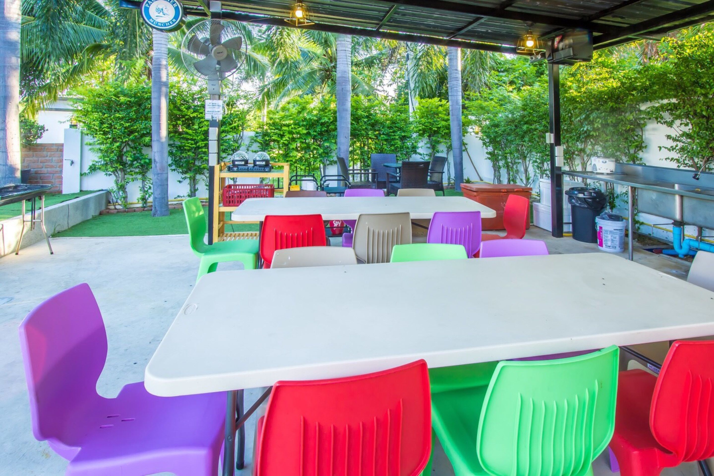 Un patio al aire libre con una mesa blanca y sillas de plástico de colores, rodeado de follaje y una barandilla de metal.