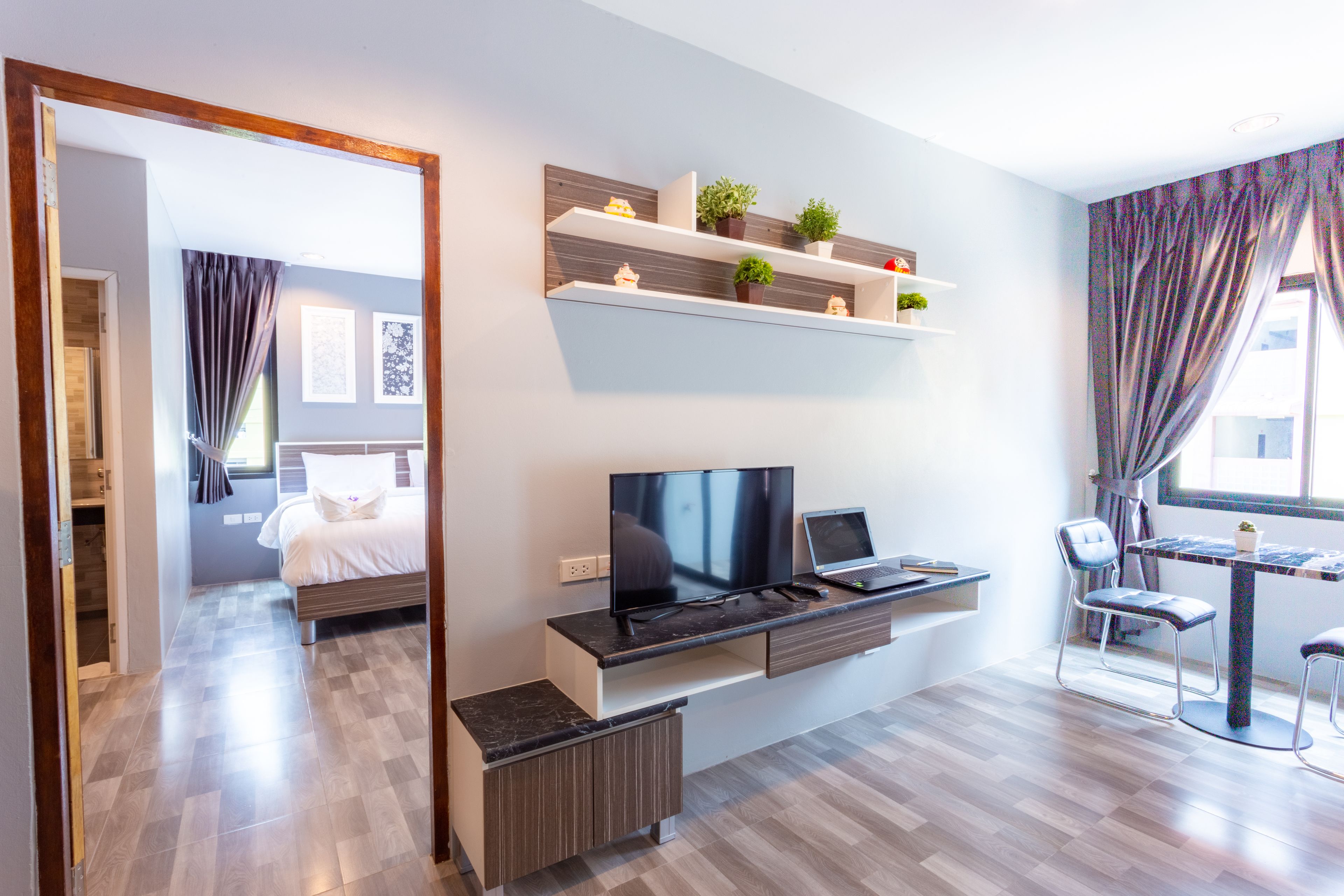 A modern hotel room with a bed, a desk with a laptop, a TV, and a dining area with chairs.