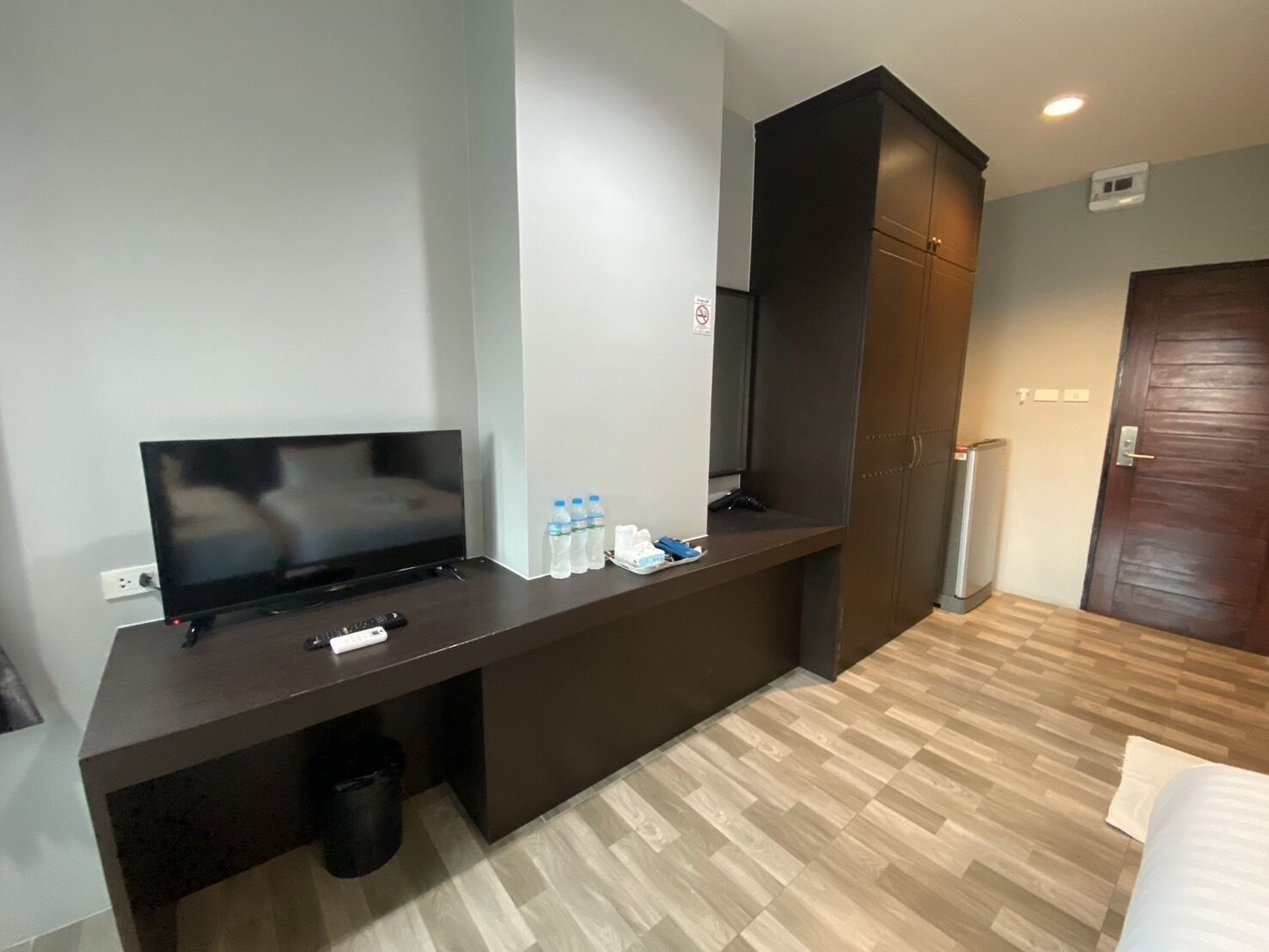 A hotel room with a flat-screen TV, a dark wooden desk, a trash bin, a water dispenser, and a wooden door.