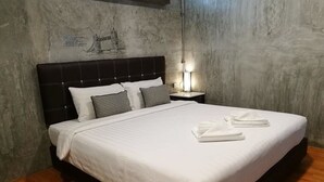 Una cama bien hecha con sábanas blancas y dos almohadas, una mesita de noche con una lámpara, y una pared decorada con un dibujo.