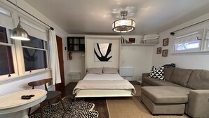 A modern bedroom with a bed, a sofa, a small table, and a chair. There is a window with curtains, a ceiling fan, and a wall-mounted shelf with books.