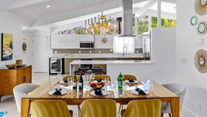 A modern kitchen with a dining area, featuring a large table set for six, a hanging chandelier, and a view of the outdoors.
