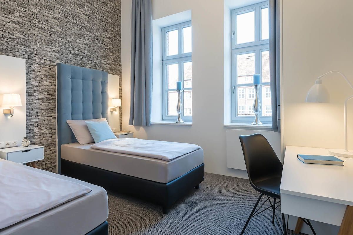 A hotel room with a bed, a desk, and a chair. There are two windows with curtains, a stone wall, and a lamp on the desk.