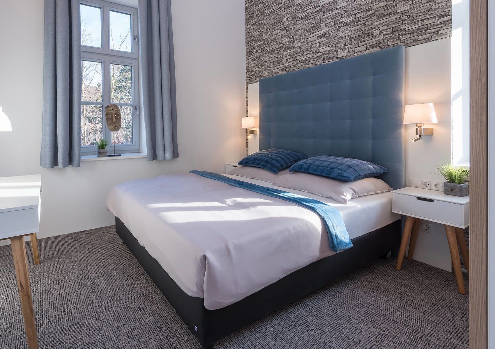 A modern bedroom with a large bed, a headboard, bedside tables, a desk, and a window with curtains.
