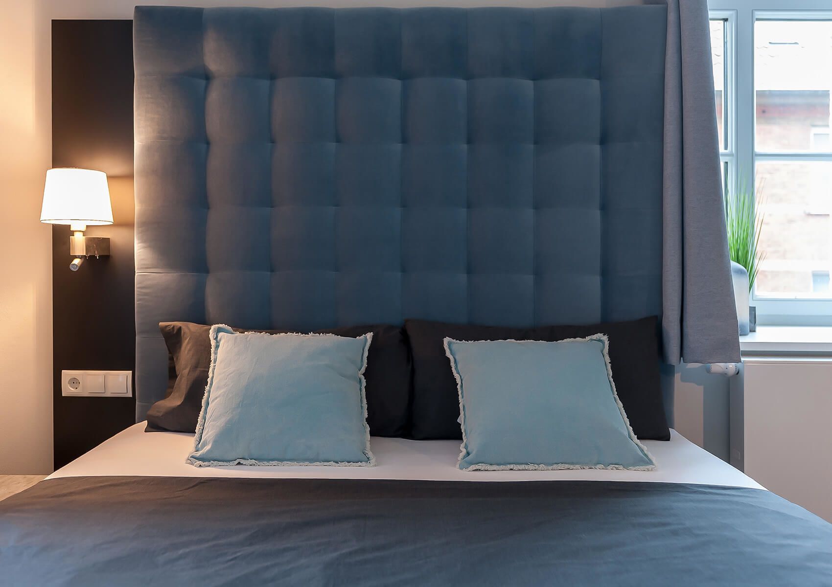 Un dormitorio con cabecera capitonada, dos almohadas, una lámpara de noche y una ventana con cortinas.