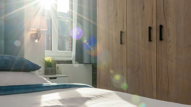 A bedroom with a bed, a wooden wardrobe, a window with sunlight, and a wall-mounted light fixture.