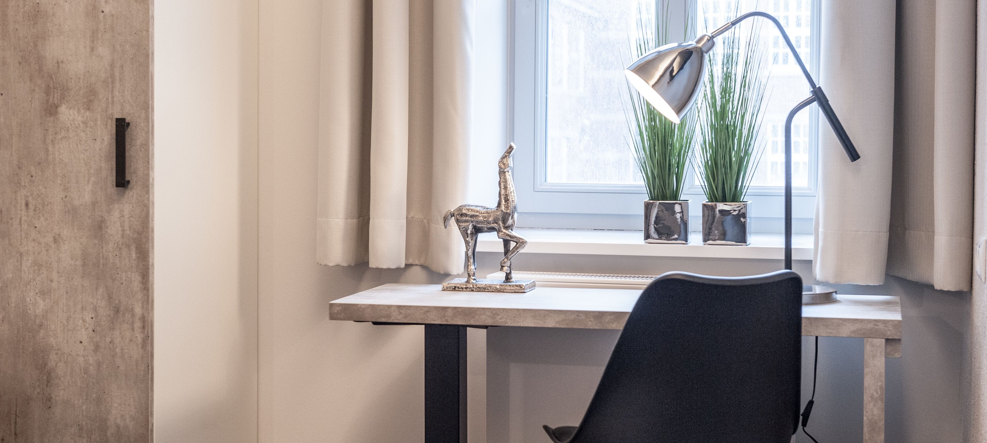 A workspace with a desk, chair, lamp, and decorative items near a window with curtains.