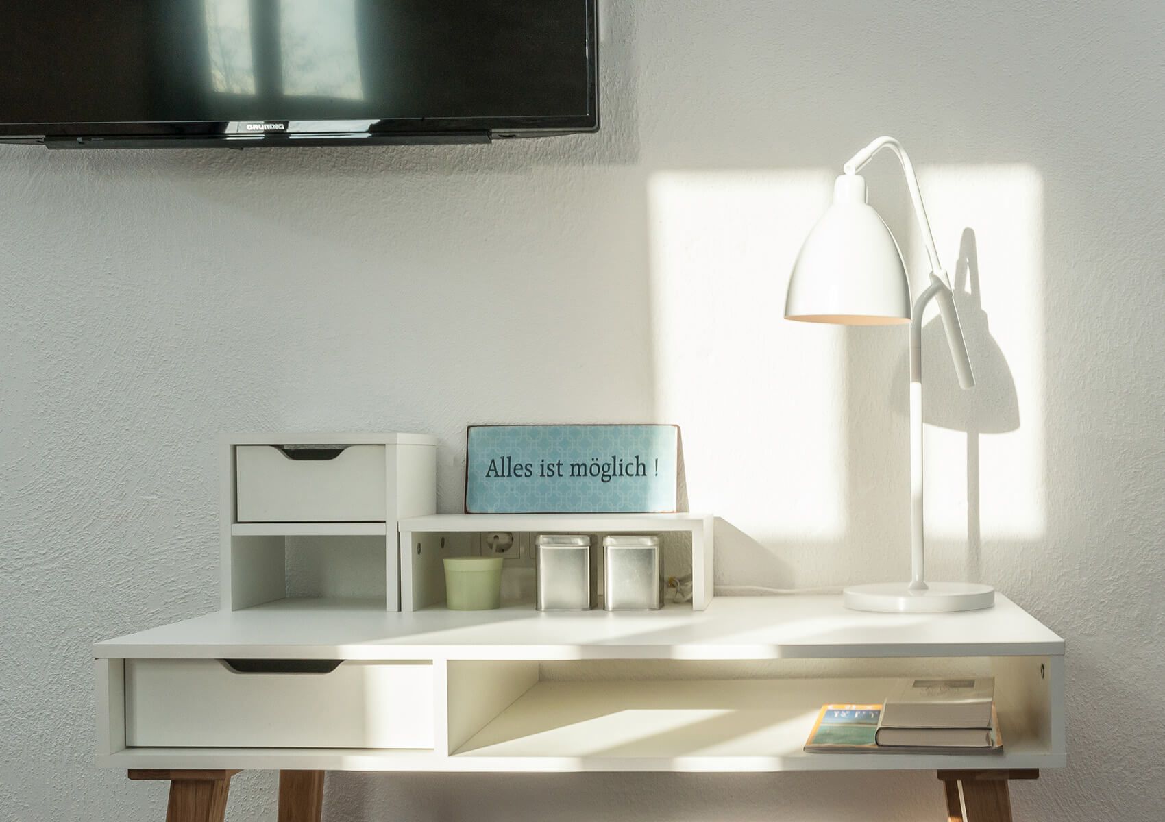 Ein weißer Schreibtisch mit einem darüber montierten Fernseher, einer weißen Schreibtischlampe, einem blauen Schild mit Text und einem Buch.