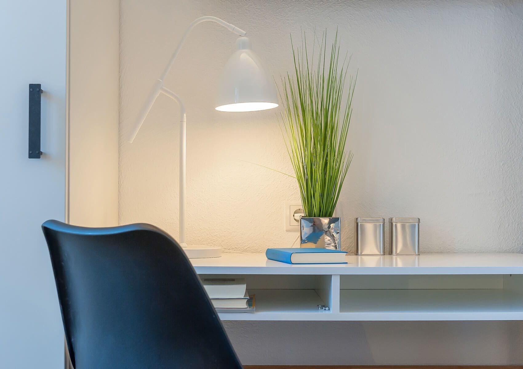 A modern desk with a white lamp, a blue book, and a plant in a reflective vase.