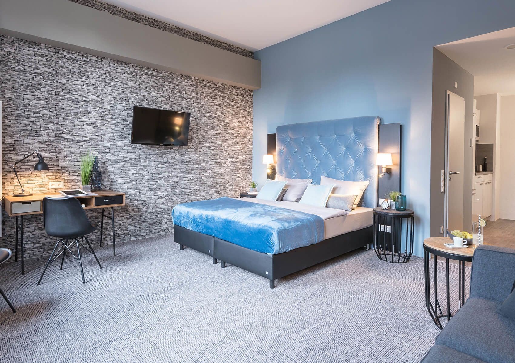A modern bedroom with a stone wall, a large bed, a desk with a chair, and a TV mounted on the wall.