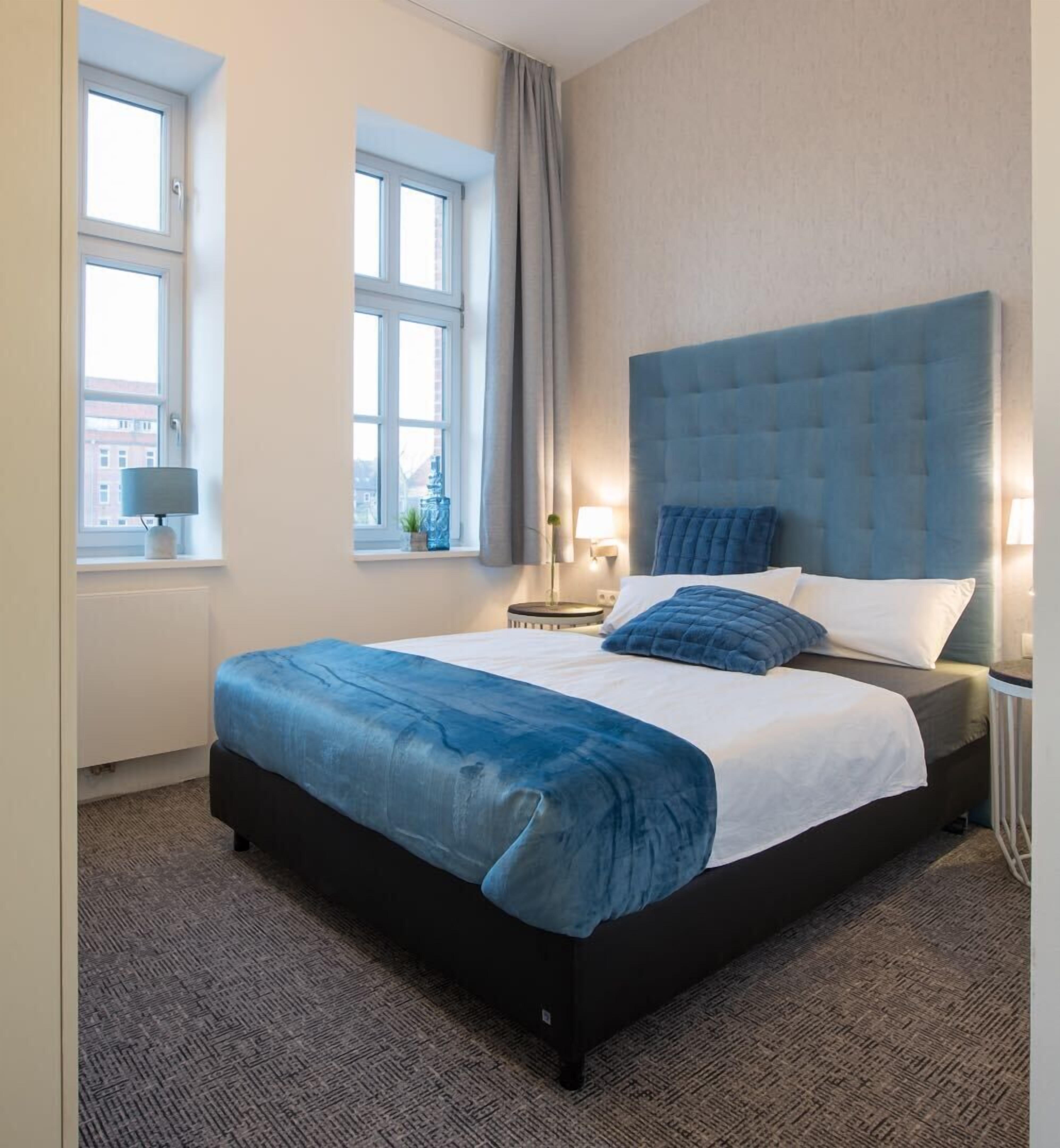 Ein Hotelzimmer mit einem großen Bett, zwei Fenstern, einem Nachttisch und einer Lampe.