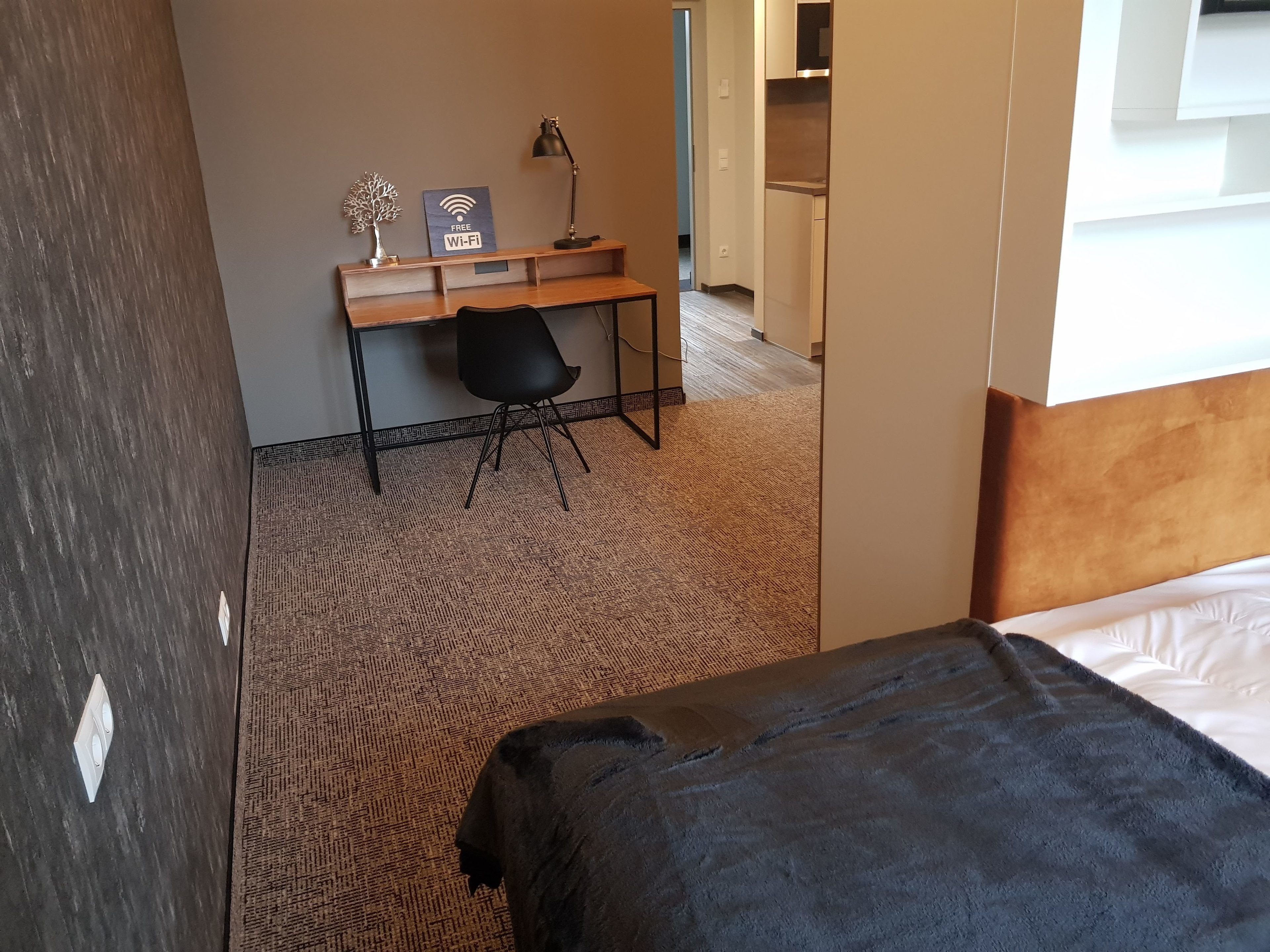 A hotel room with a bed, a desk with a chair, a lamp, and a sign indicating free Wi-Fi.