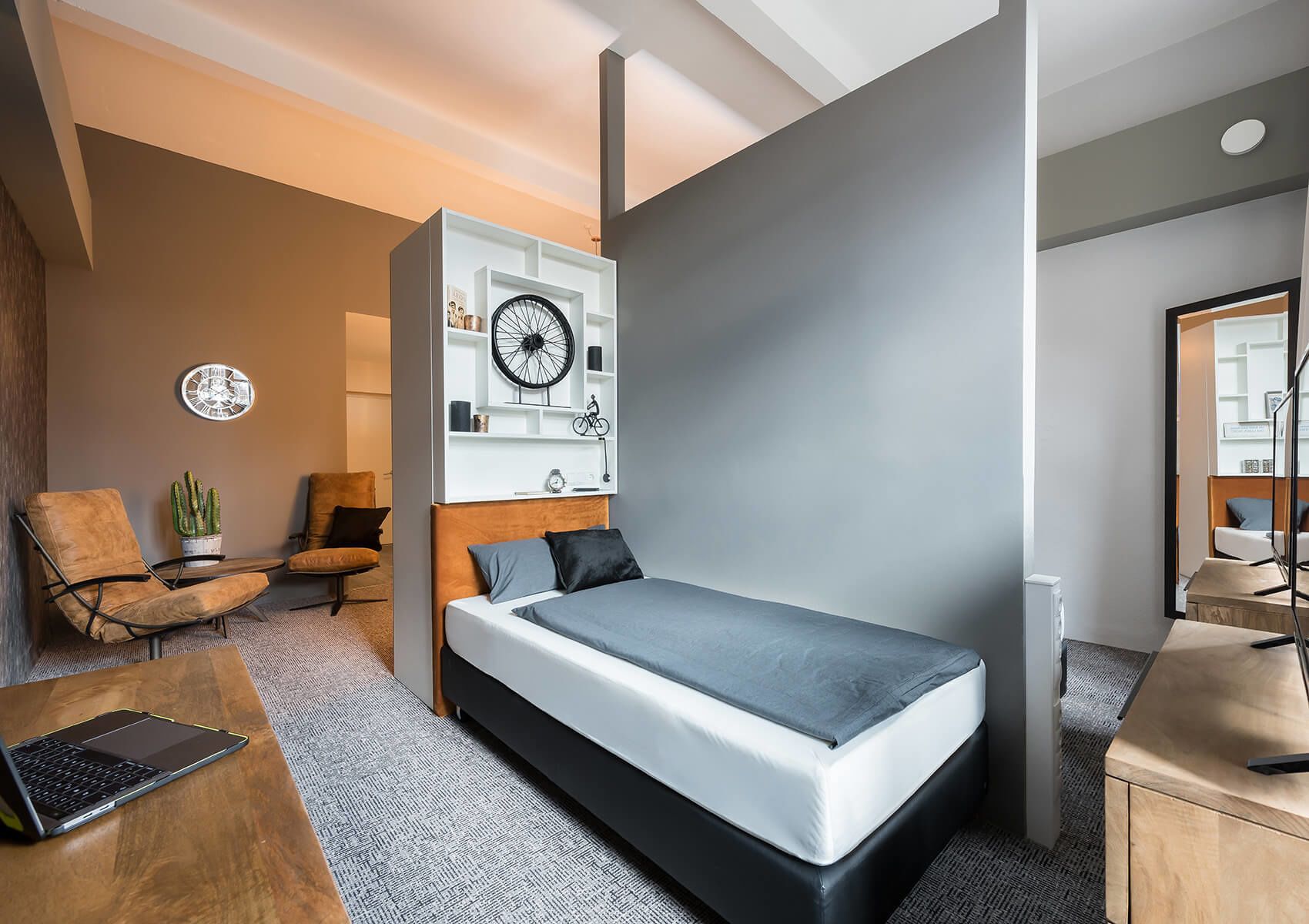 A modern bedroom with a bed, a desk with a laptop, a chair, and a wall-mounted shelf with a bicycle.