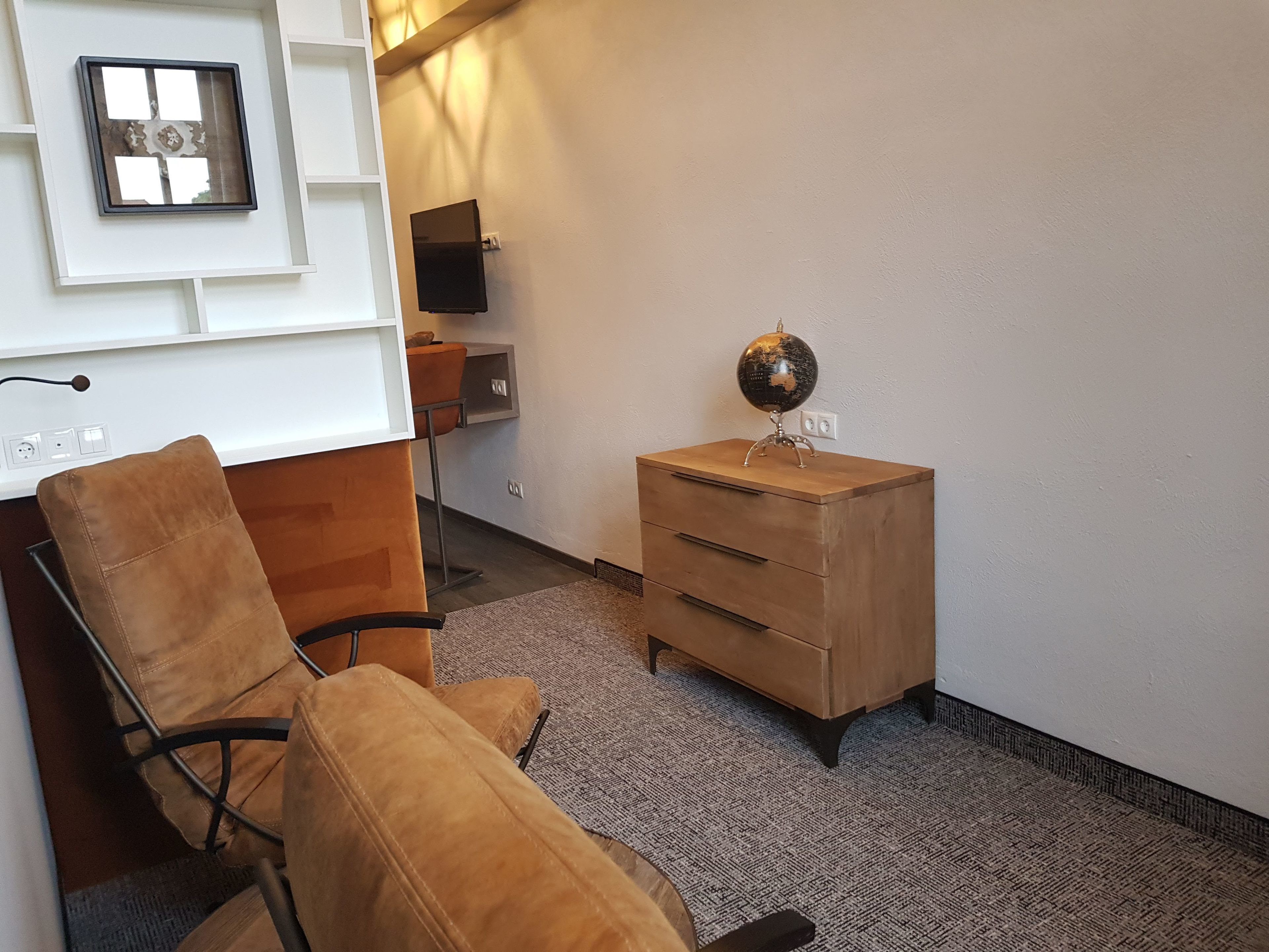 A hotel room with a wooden desk, a chair, a globe on a small wooden cabinet, and a framed picture on the wall.
