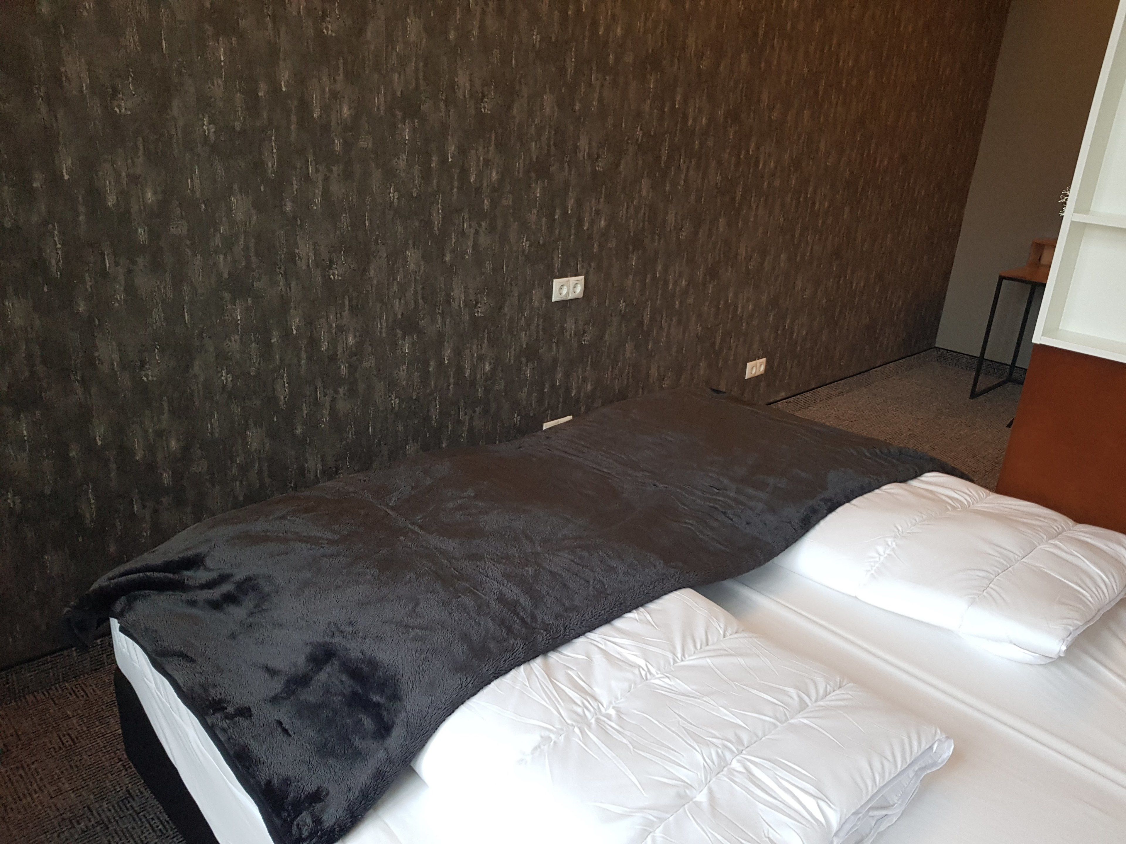 A hotel room with a bed, a dark textured wall, and a small table with a chair.