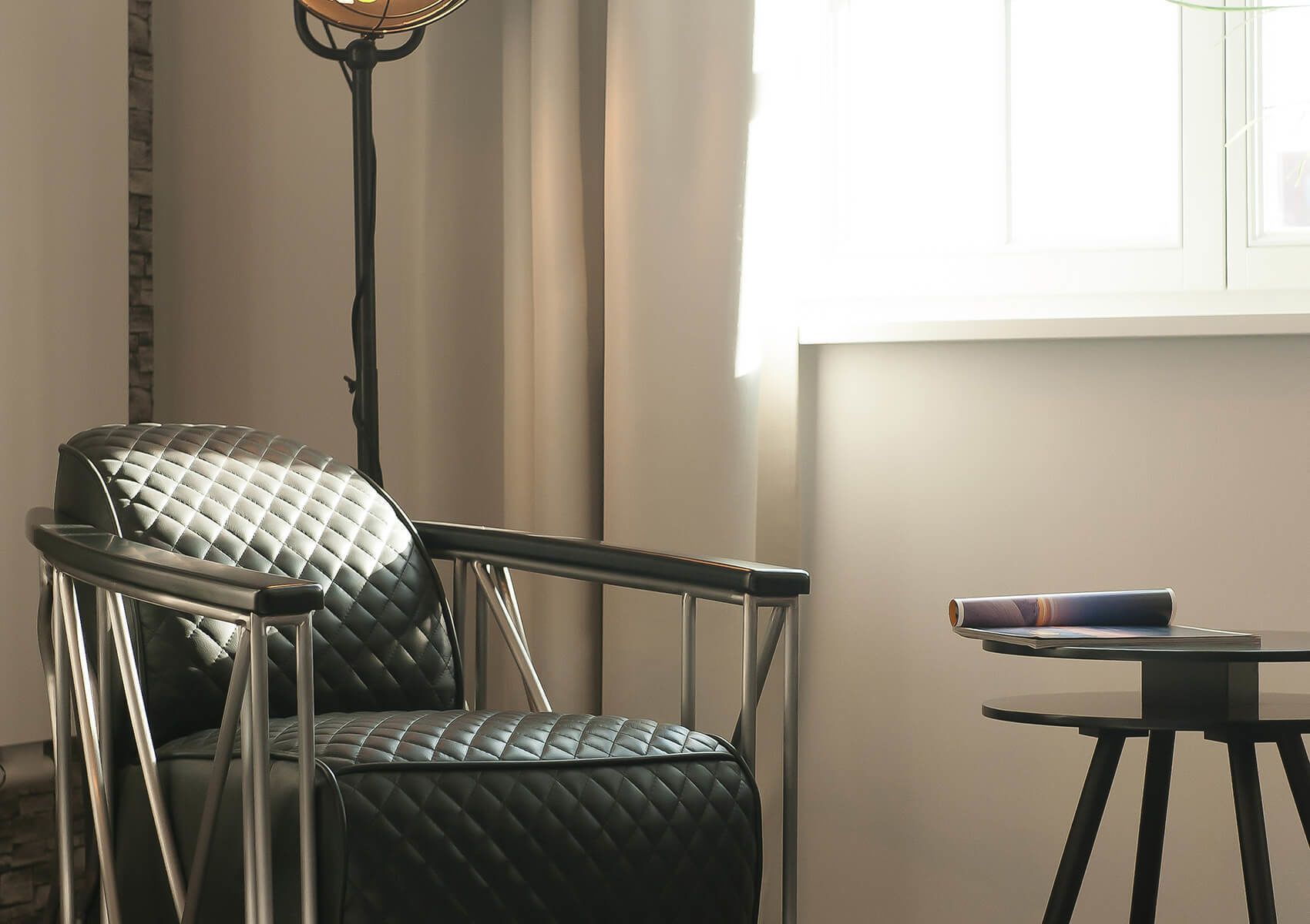 Een moderne kamer met een zwarte leren fauteuil, een vloerlamp, een klein tafeltje met een boek en een raam met gordijnen.