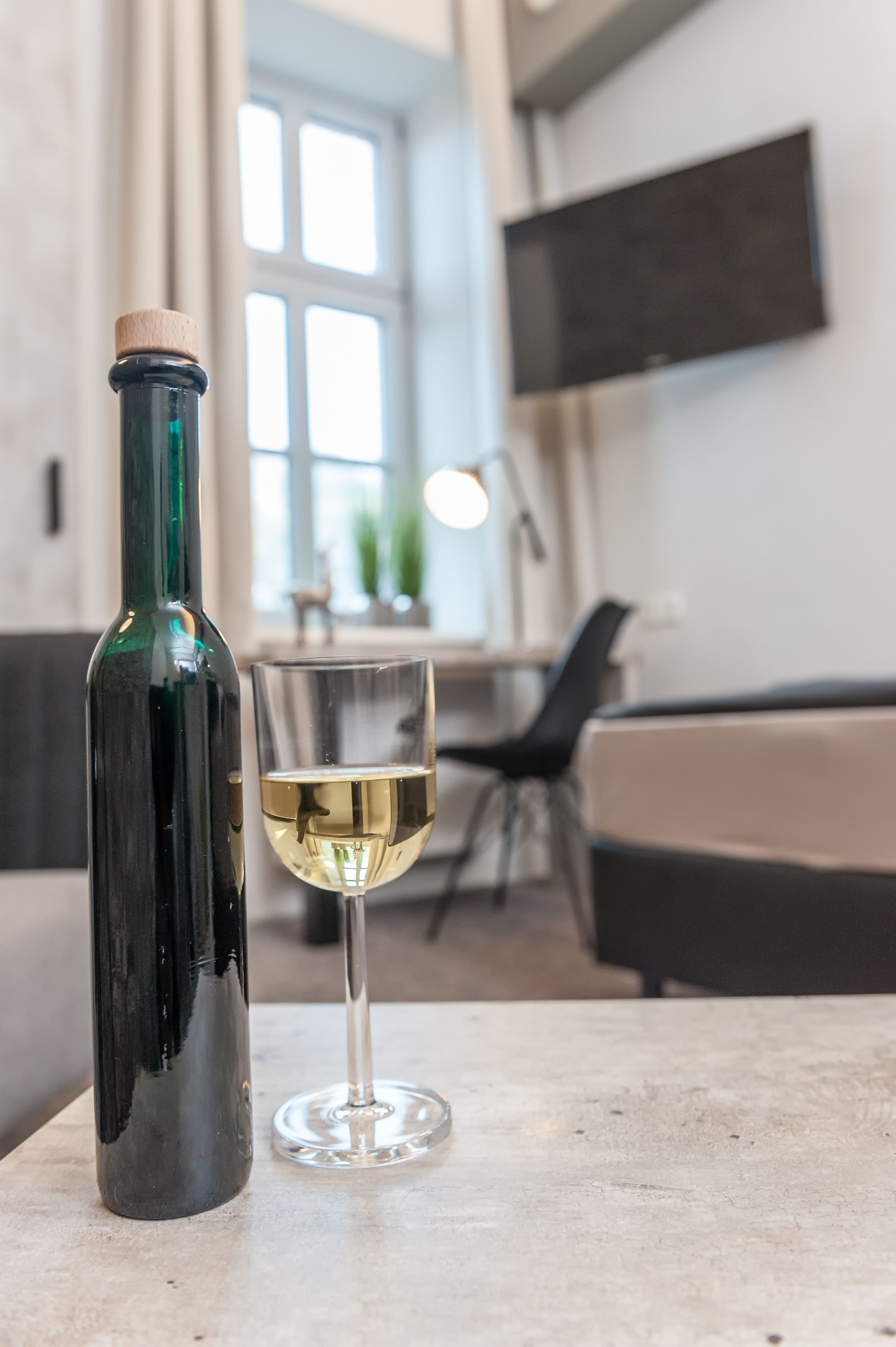 A bottle of wine and a glass on a table in a modern kitchen with a large window and a flat-screen TV mounted on the wall.
