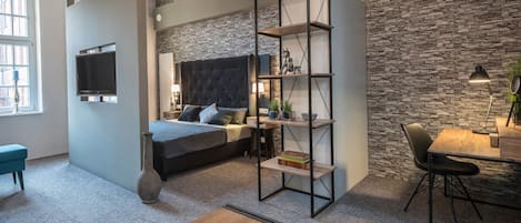 Una sala de estar moderna con una pared de piedra, una cama grande con cabecera capitonada, una mesa de centro de madera, un televisor montado en la pared y un comedor con mesa y sillas.