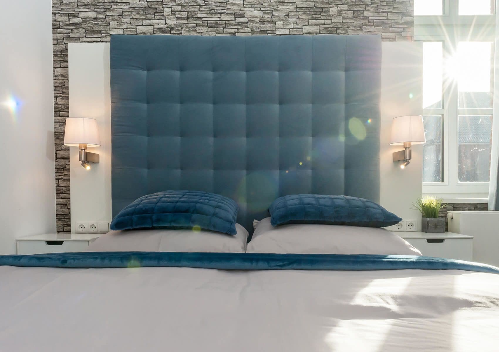 A bedroom with a stone wall, a tufted headboard, two pillows, and bedside lamps.