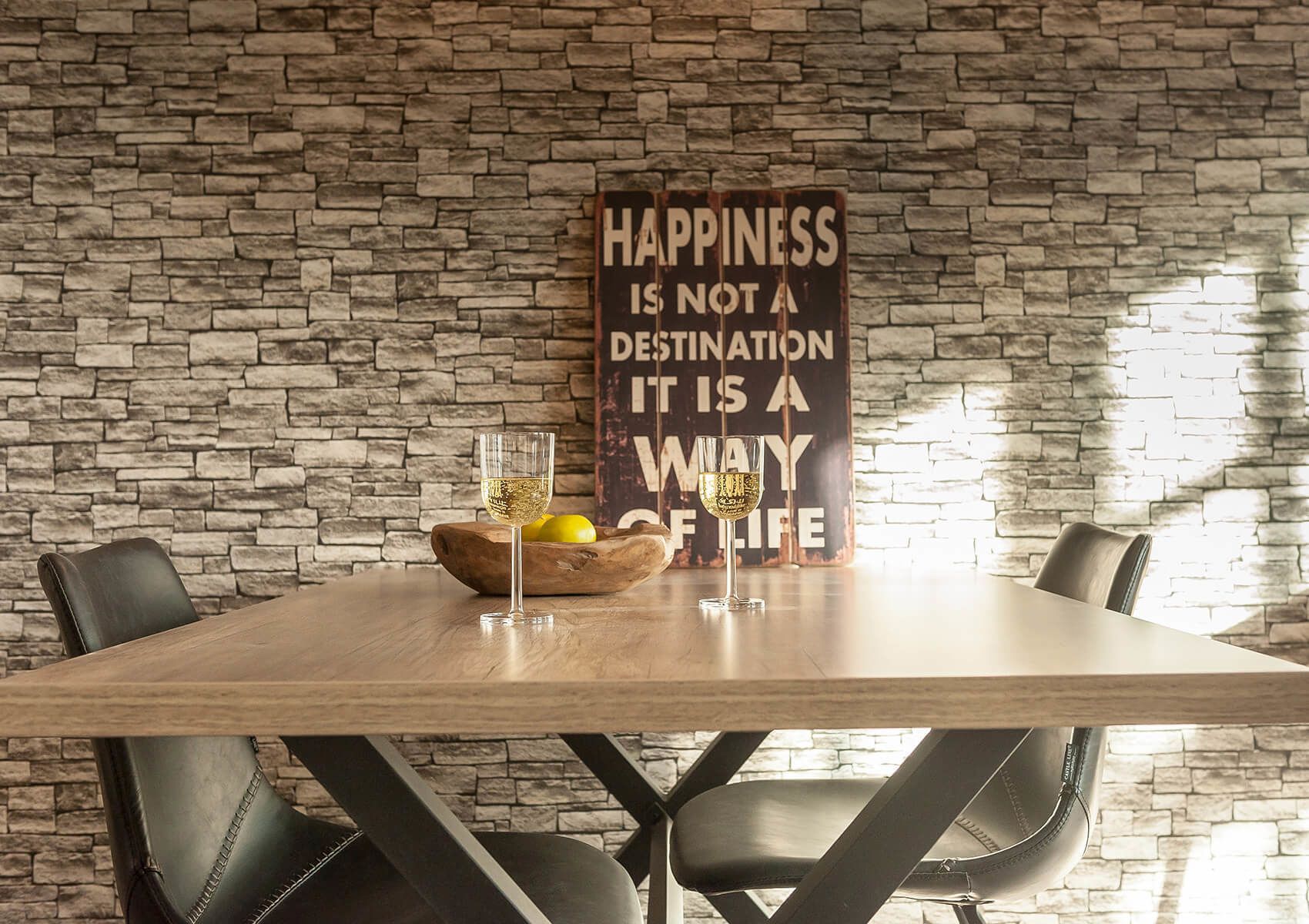 A dining table with two glasses of wine and a bowl of lemons, set against a stone wall with a motivational sign.