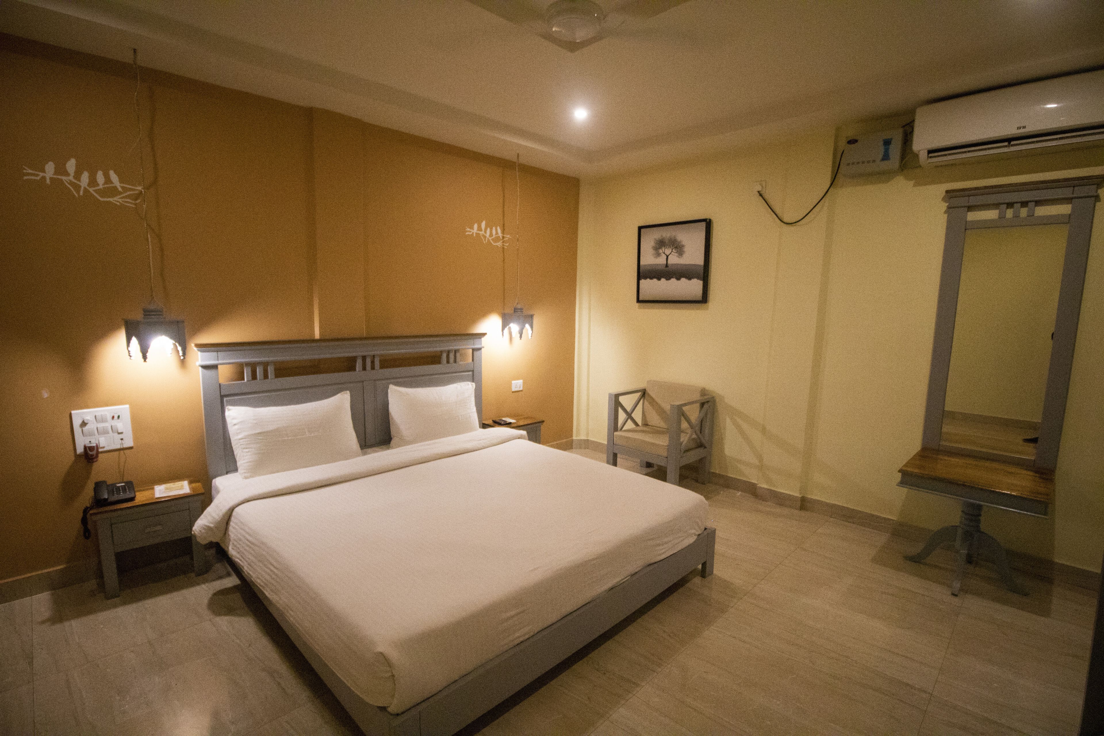 Une chambre d’hôtel avec un grand lit, deux tables de chevet, une petite table, une chaise, un climatiseur fixé au mur et un tableau encadré.