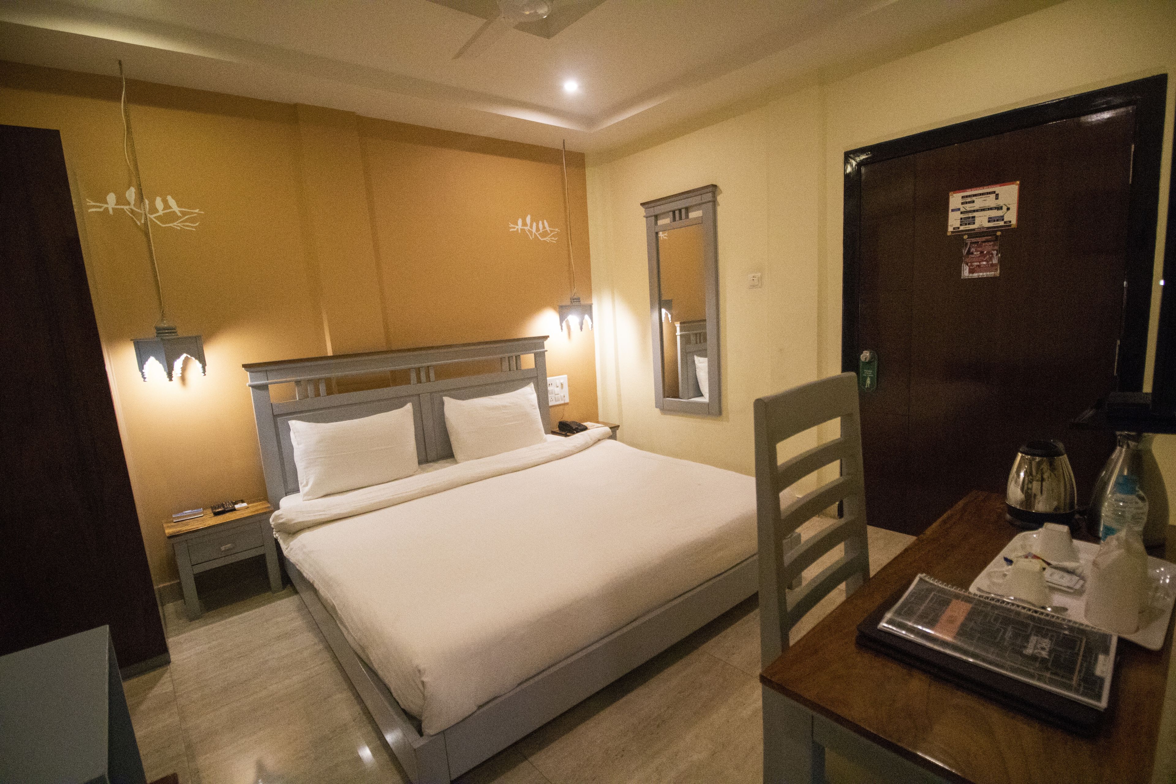 A hotel room with a bed, bedside table, lamp, mirror, and a desk with a keyboard and coffee maker.
